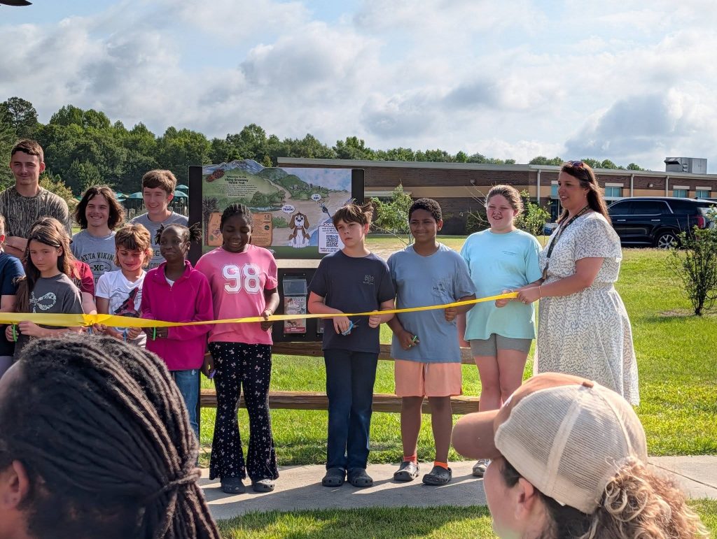 A group of children and an adult stand outside holding a yellow ribbon for a ribbon-cutting ceremony, with a sign and school building in the background.