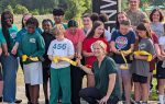 A group of children and an adult smile and cut a yellow ribbon outdoors, participating in a ribbon-cutting ceremony on a sunny day.