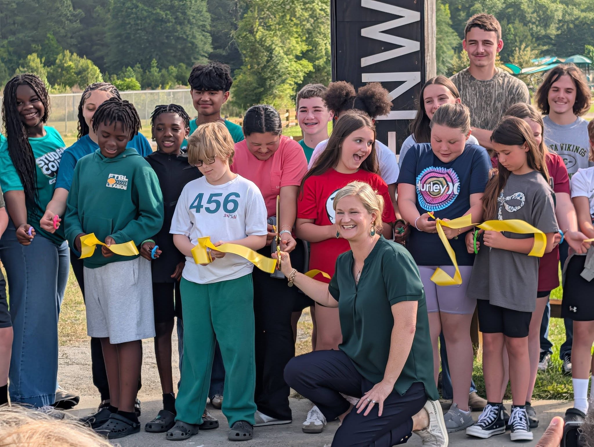 A group of children and an adult smile and cut a yellow ribbon outdoors, participating in a ribbon-cutting ceremony on a sunny day.