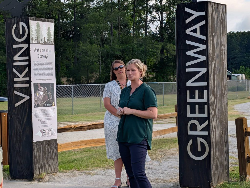 Two women stand between large wooden signs reading "Viking Greenway" at an outdoor trail entrance, with trees and grass visible in the background.
