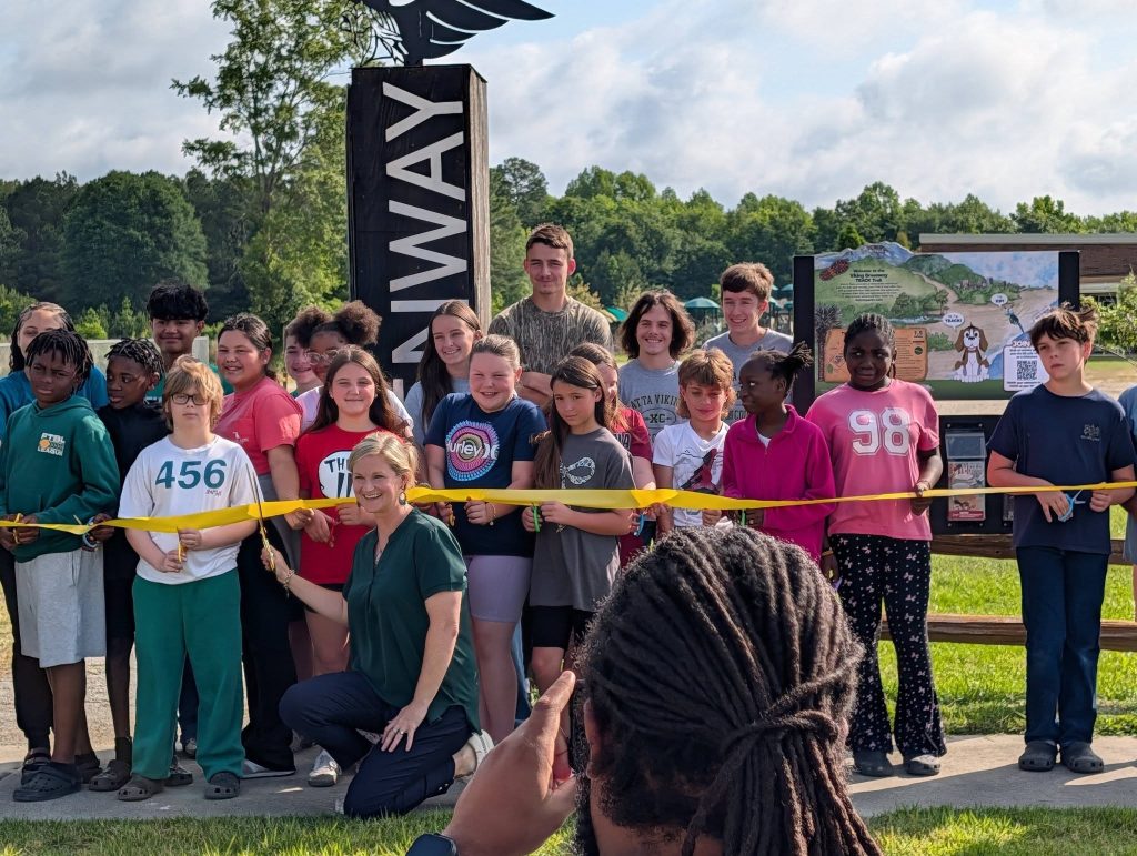 A group of children and an adult pose for a ribbon-cutting ceremony outdoors near a sign that reads "RUNWAY" with trees and a map in the background.
