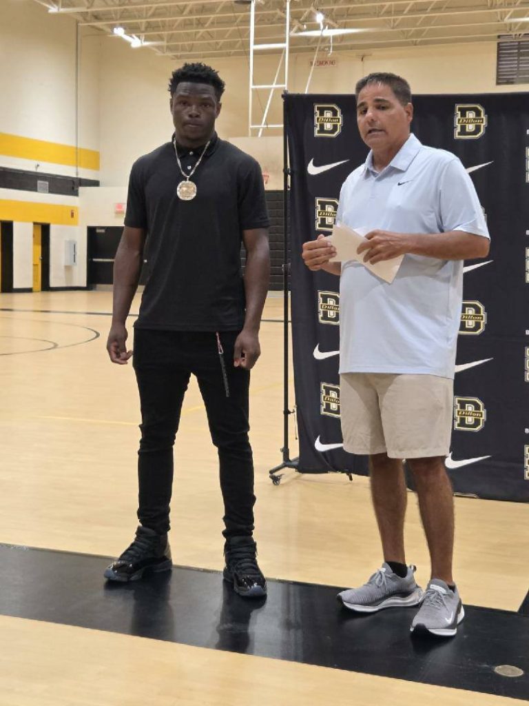 Two men standing in a gymnasium; one wearing all black with a medallion, the other in a light shirt and shorts, holding papers.