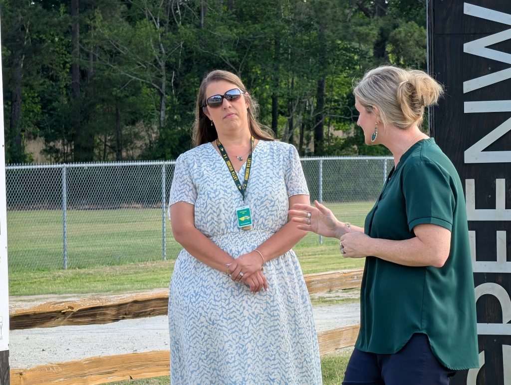 Two women stand outside near a fence, one wearing sunglasses and a name badge, listening as the other gestures and speaks, with trees and a sign in the background.