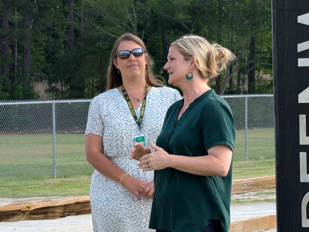 Two women stand outdoors near a fence, one wearing sunglasses and a badge, while the other speaks, with trees in the background.