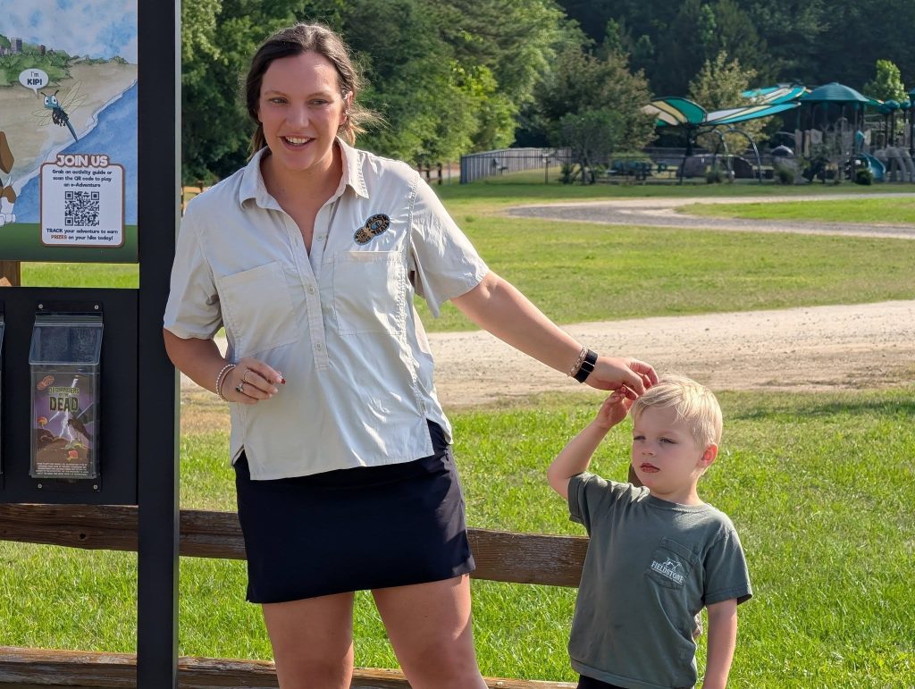 Woman and young boy standing outdoors by an information board, with a playground and trees in the background on a sunny day.