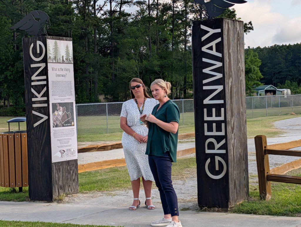 Two women stand between large wooden signs reading "Viking Greenway" at an outdoor park with trees and a chain-link fence in the background.