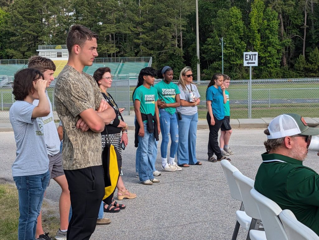 A group of students and adults stand outside near a sports field, some wearing "Student Council" shirts, listening attentively. Trees and a stadium are visible in the background.