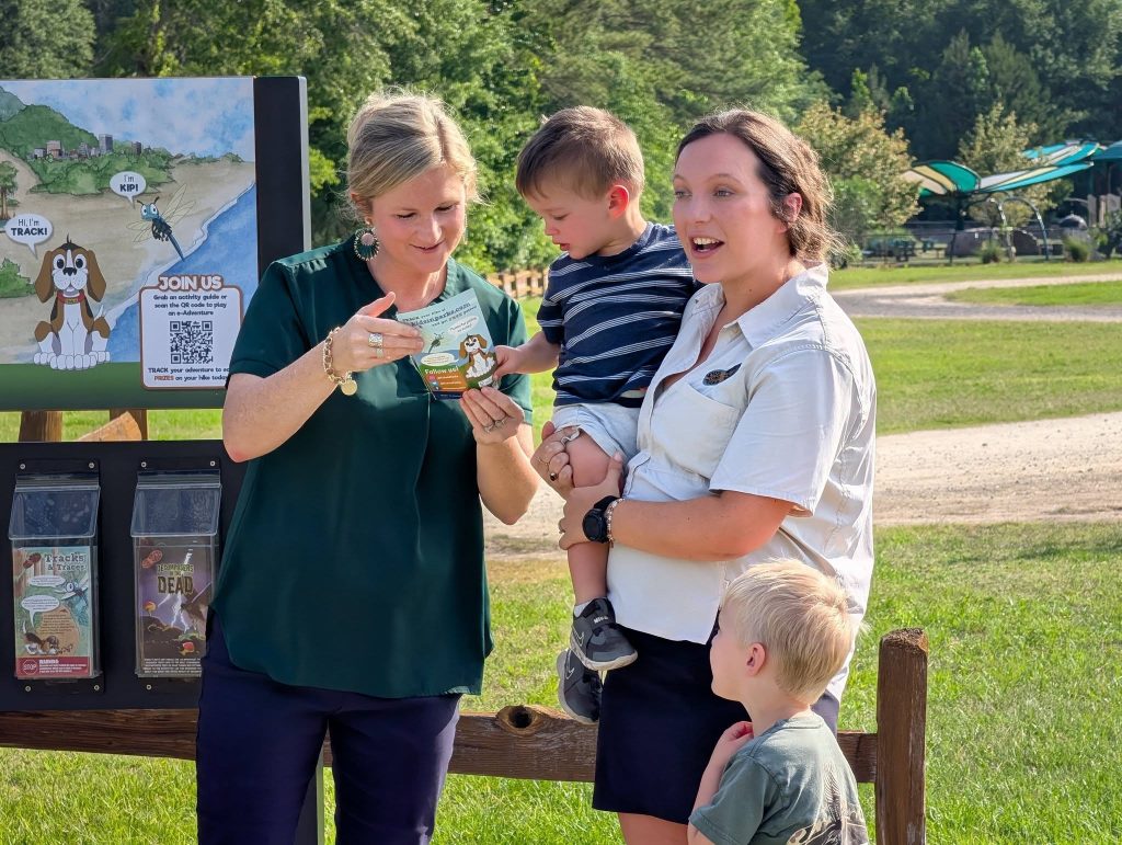 Two women and two young boys look at a brochure outdoors near an informational sign in a park with trees and grassy area in the background.