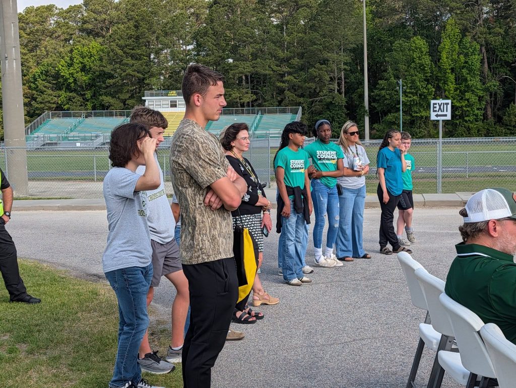 A group of people, including students wearing "Student Council" shirts, stand outdoors near bleachers and an "EXIT" sign, listening attentively.