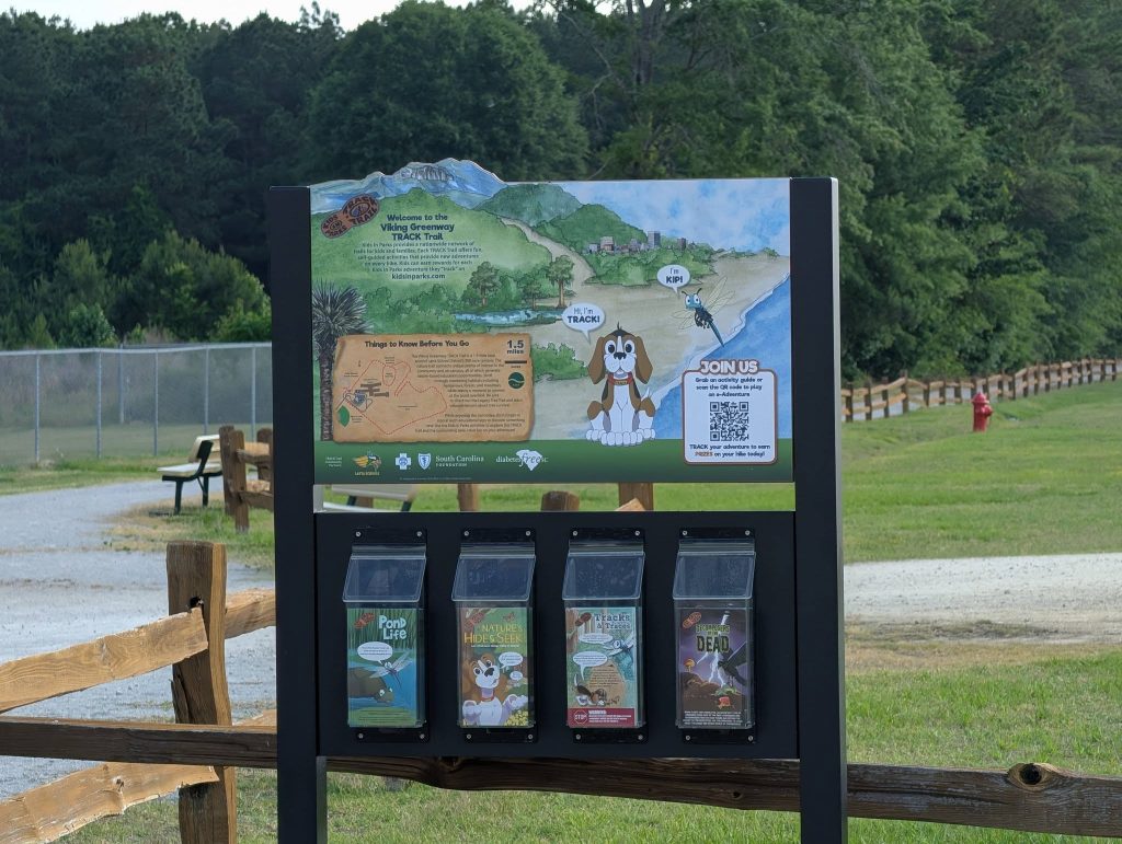 Outdoor informational sign at the Viking Greenway TRACK Trail with illustrated map, cartoon dog, trail guides, and pamphlet dispensers, surrounded by trees and fenced path.