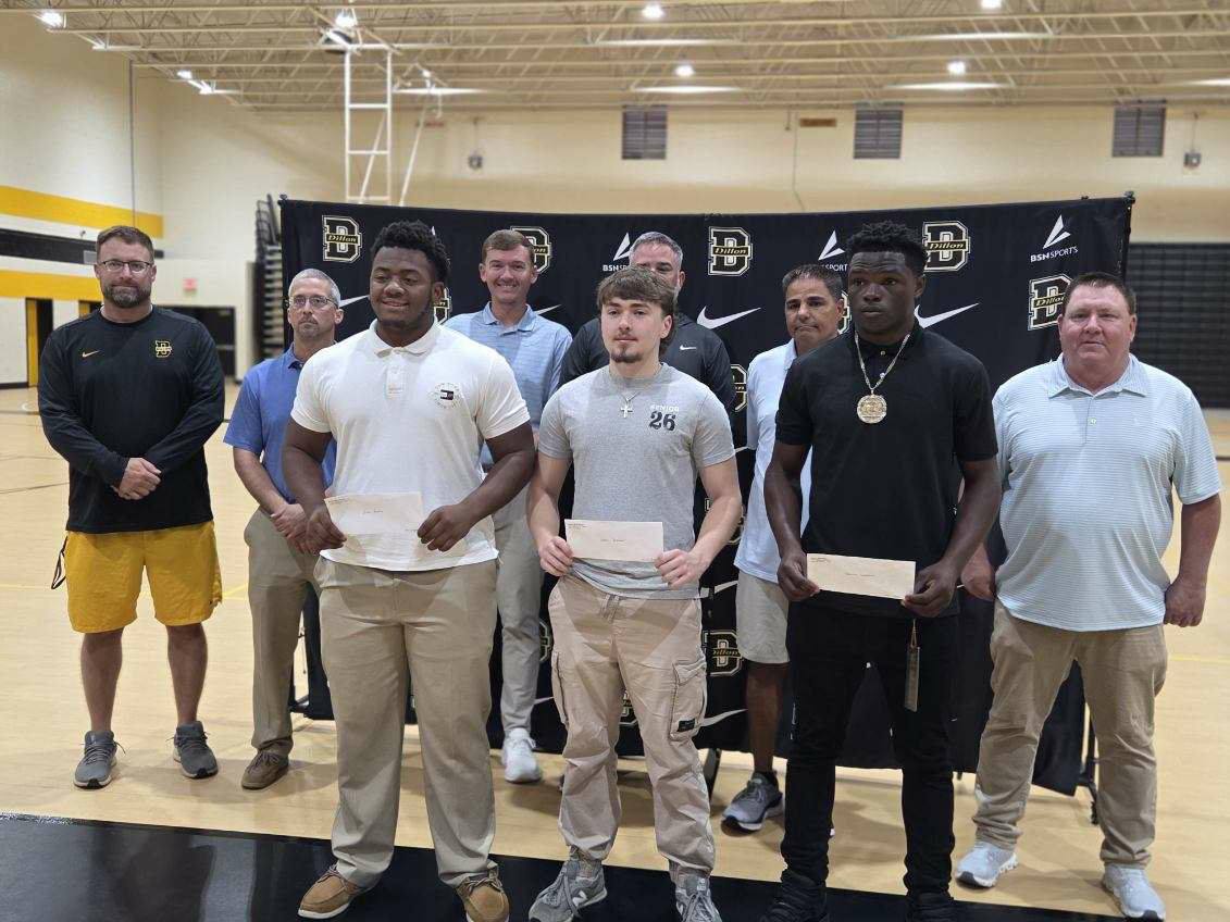 A group of young men and adults pose indoors, with three holding envelopes, in front of a sports-themed backdrop at a gymnasium.