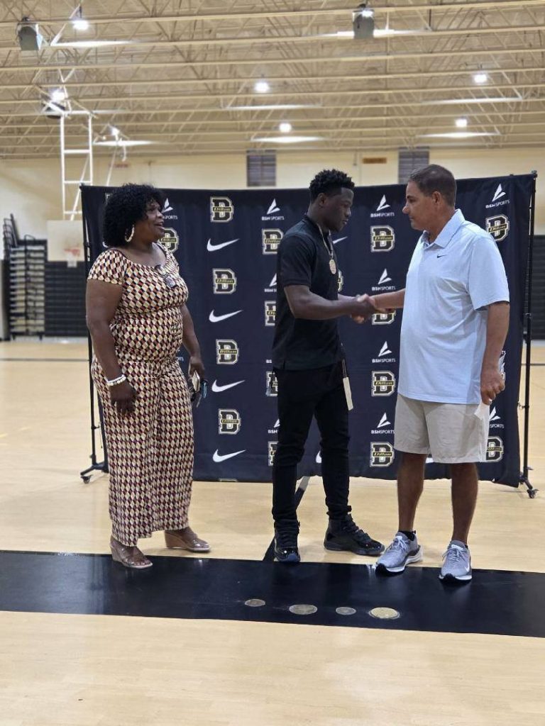 Three people are in a gymnasium; two men shake hands while a woman stands nearby, with a school-branded backdrop behind them.