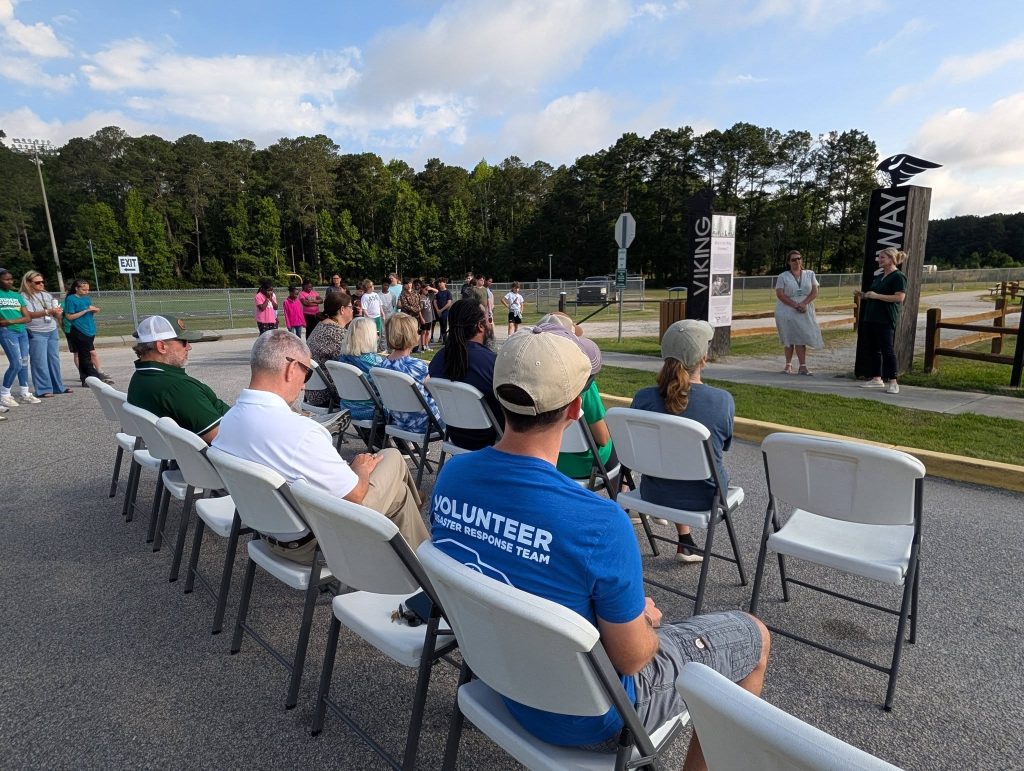 People seated in folding chairs outdoors, listening to two speakers near a sign labeled "Viking Way." Forest and sports field visible in the background, partly cloudy sky.