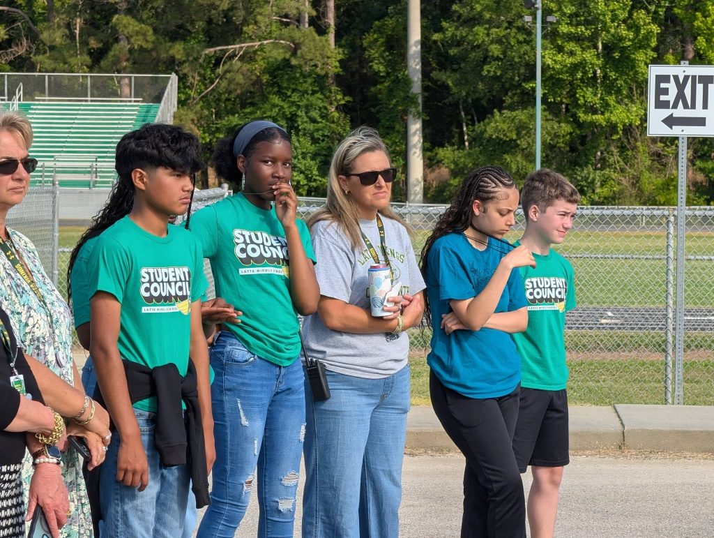 A group of students and an adult stand outdoors near a fence and "EXIT" sign, several wearing "Student Council" shirts, looking ahead seriously.
