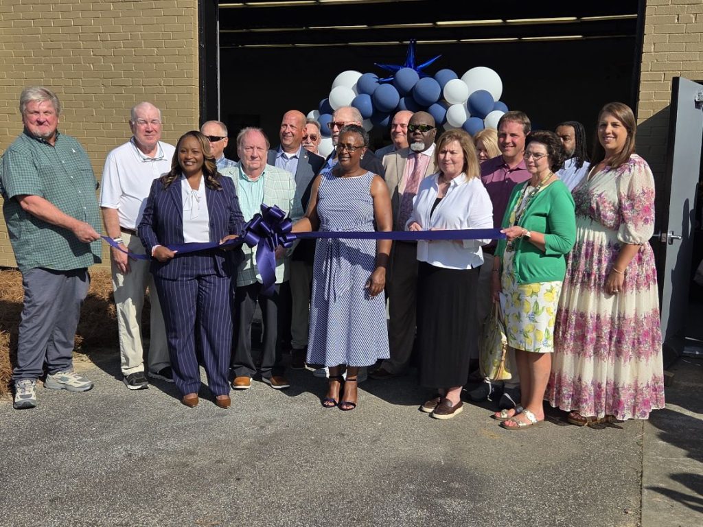 A group of people stand outside a building for a ribbon-cutting ceremony, with blue and white balloons decorating the entrance.