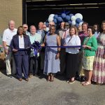 A group of people stand outside a building for a ribbon-cutting ceremony, with blue and white balloons decorating the entrance.