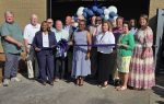 A group of people stand outside a building for a ribbon-cutting ceremony, with blue and white balloons decorating the entrance.