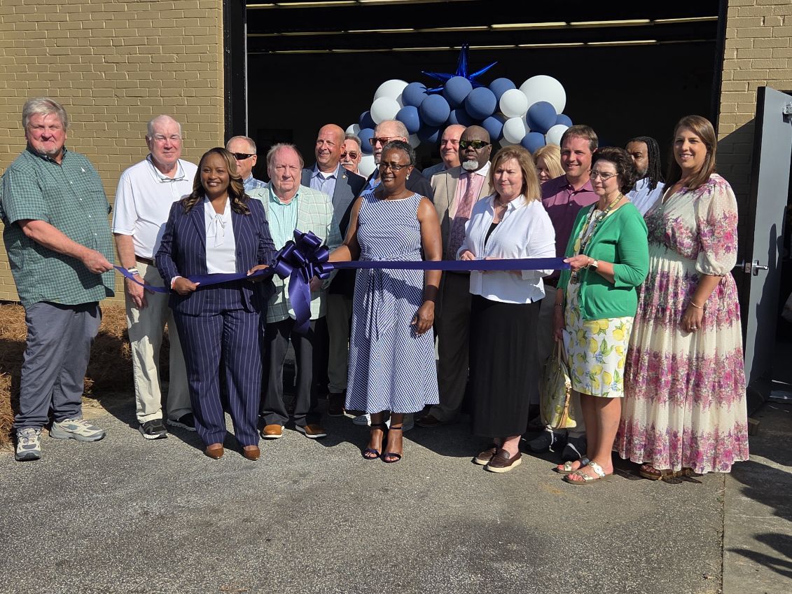 A group of people stand outside a building for a ribbon-cutting ceremony, with blue and white balloons decorating the entrance.