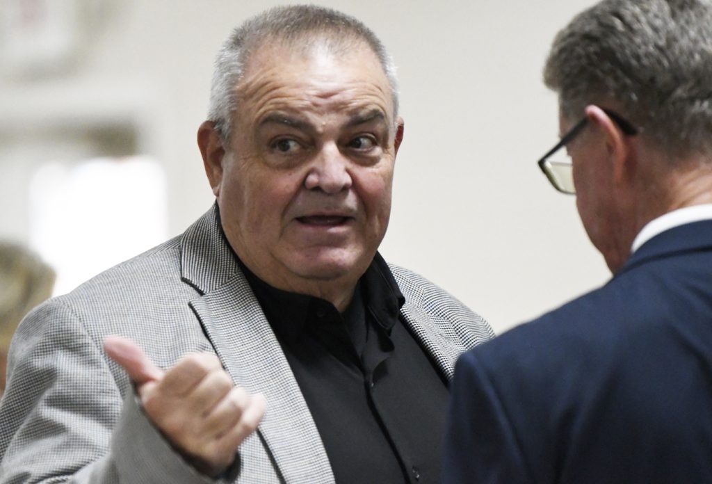 older man in a gray blazer talking and gesturing with his thumb up while speaking to another man in a suit.