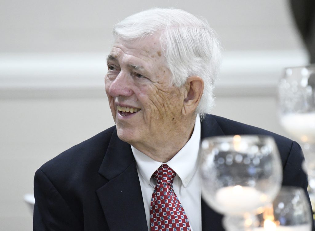 Elderly man with white hair in a suit and red patterned tie smiling at an indoor event, with glass candle holders visible in the foreground.