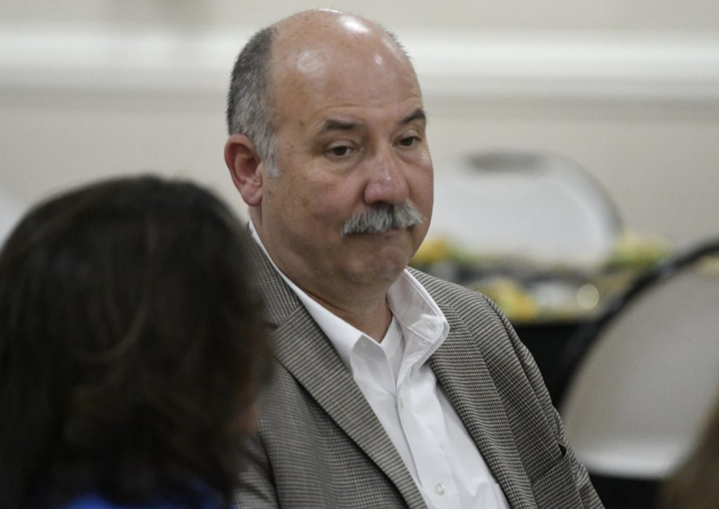 Bald man with a mustache wearing a gray suit jacket and white shirt, seated indoors at an event, looking forward.