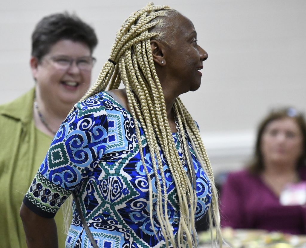 Woman with long blonde braids and blue patterned dress stands indoors, smiling, with two other women visible in background, one wearing glasses and green.