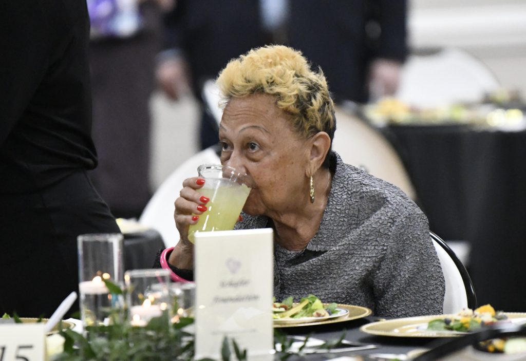 older woman with short blonde hair sips a yellow drink at a formal event, seated at a decorated table with plates of food.