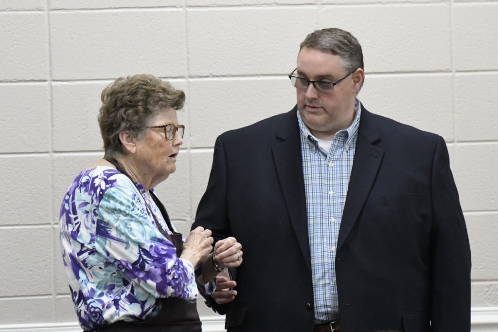 elderly woman in a floral blouse talks to a man in a suit jacket against a neutral wall background.