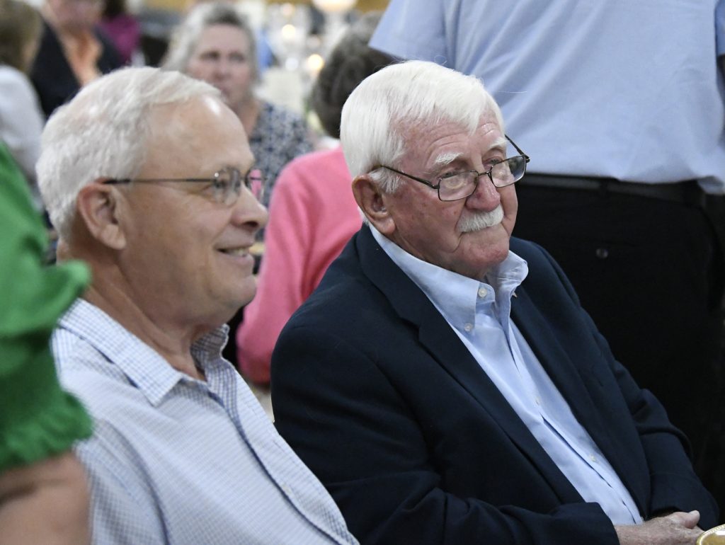 Two older men with glasses sit side by side at an indoor event, surrounded by people in the background.