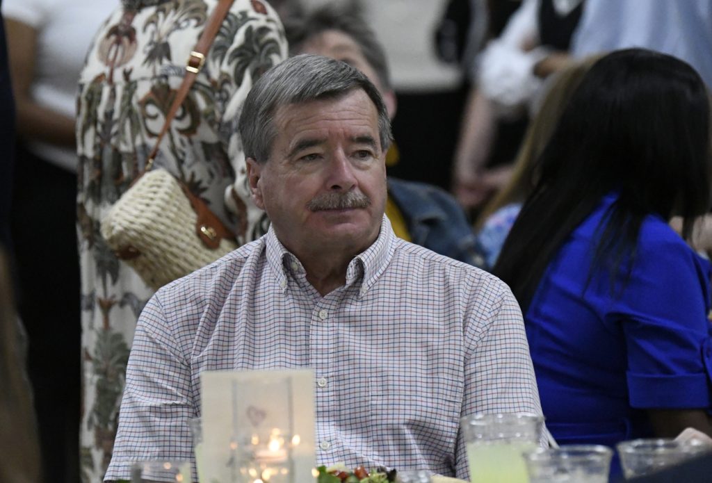 older man in a checkered shirt sits at a table with candles and food, surrounded by people at an indoor event.