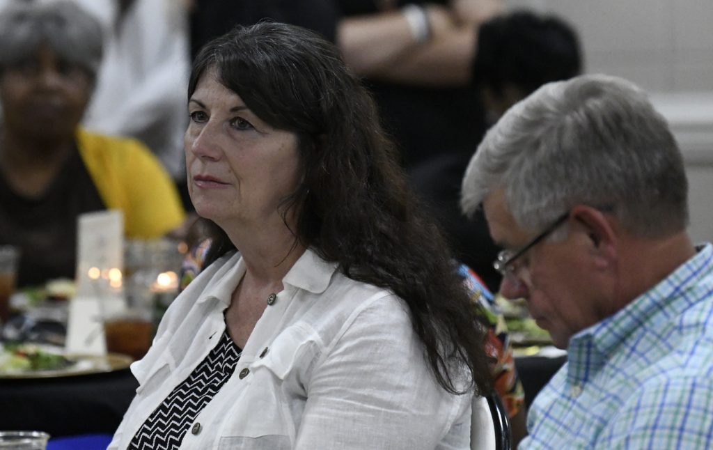 A woman and a man sit at a table during an indoor event, listening attentively among a group of people.