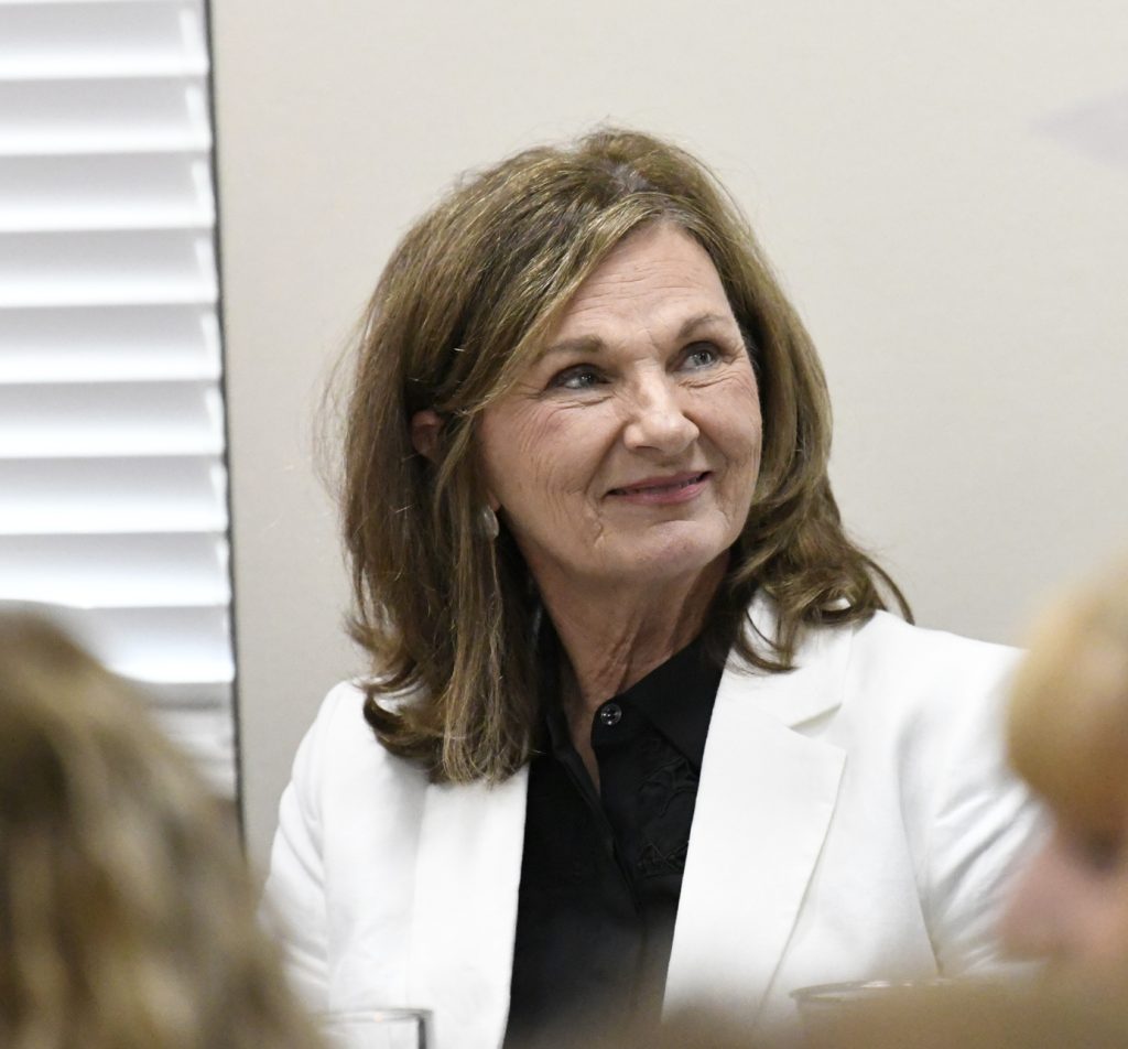A woman with shoulder-length brown hair wearing a white blazer and black blouse, smiling and looking to the side, sits indoors near a window with blinds.