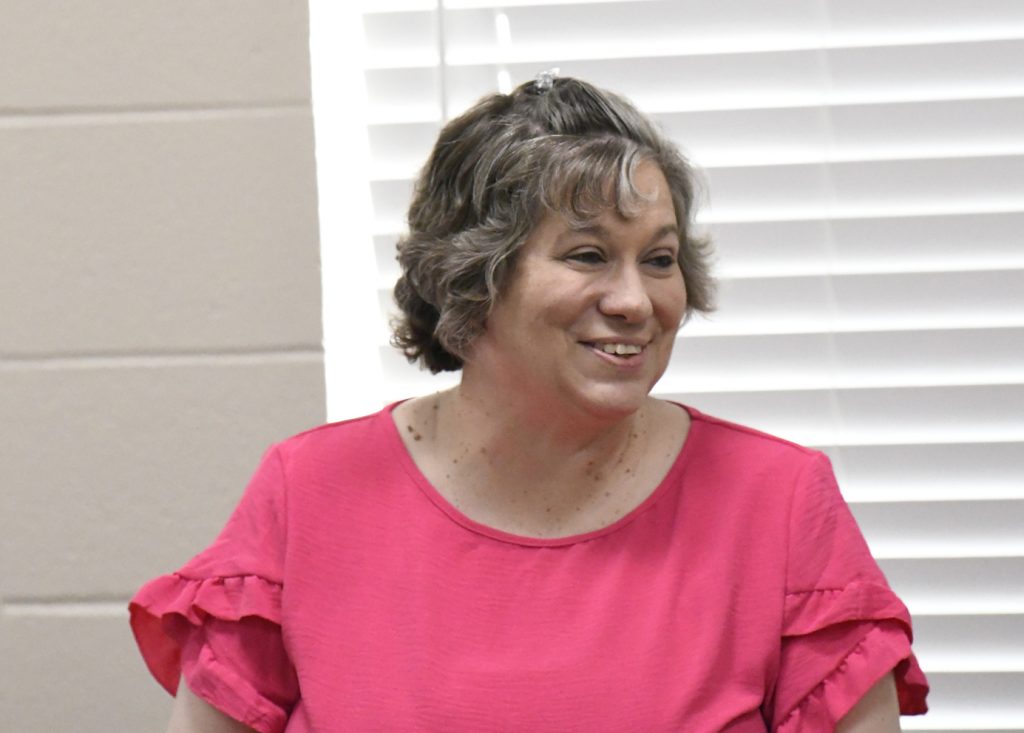 A woman with short, wavy gray hair wearing a bright pink top smiles in front of a window with white blinds.