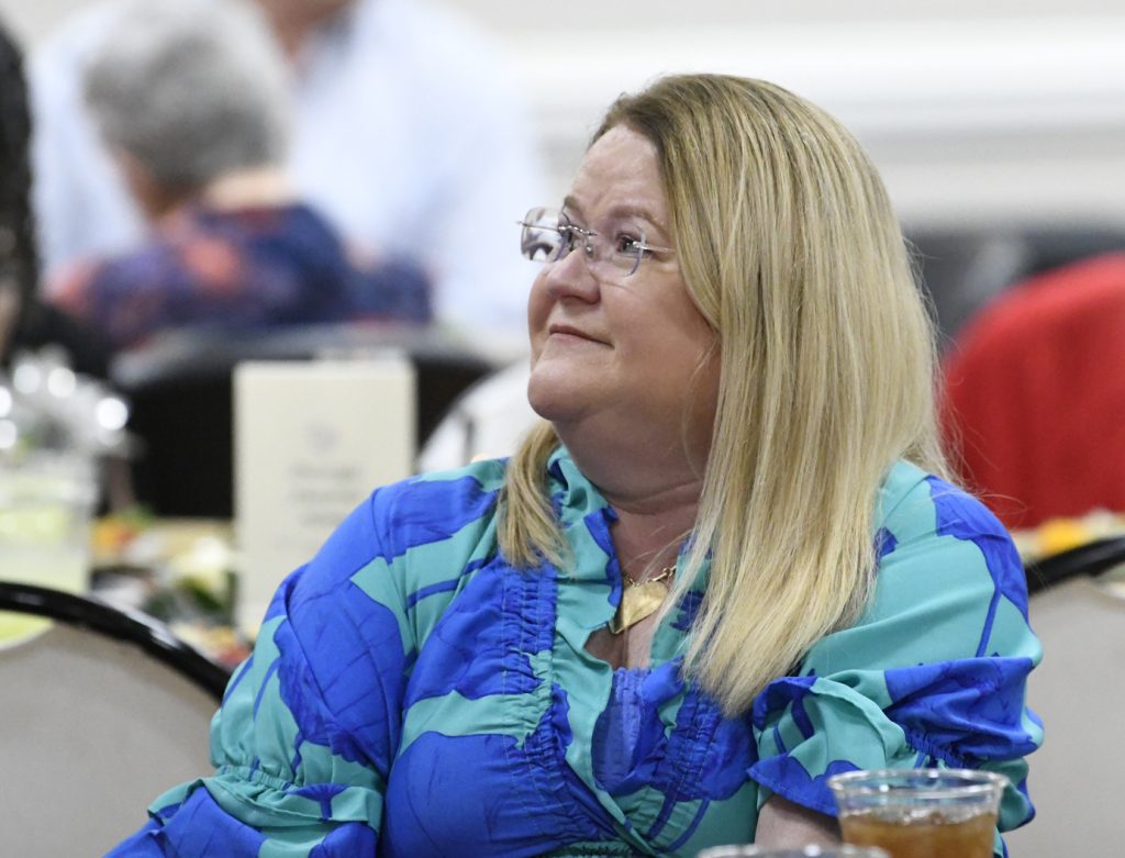 A woman with long blonde hair and glasses wearing a blue patterned blouse sits at a table, looking to the side in a crowded indoor setting.