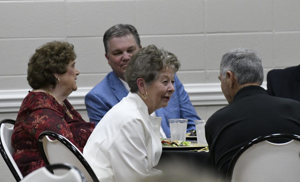 Four older adults are seated at a table, engaged in conversation during a formal meal, with food and drinks in front of them.