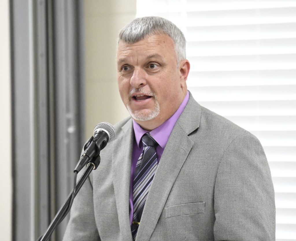 A man in a gray suit and purple shirt speaks into a microphone indoors, standing in front of white blinds.