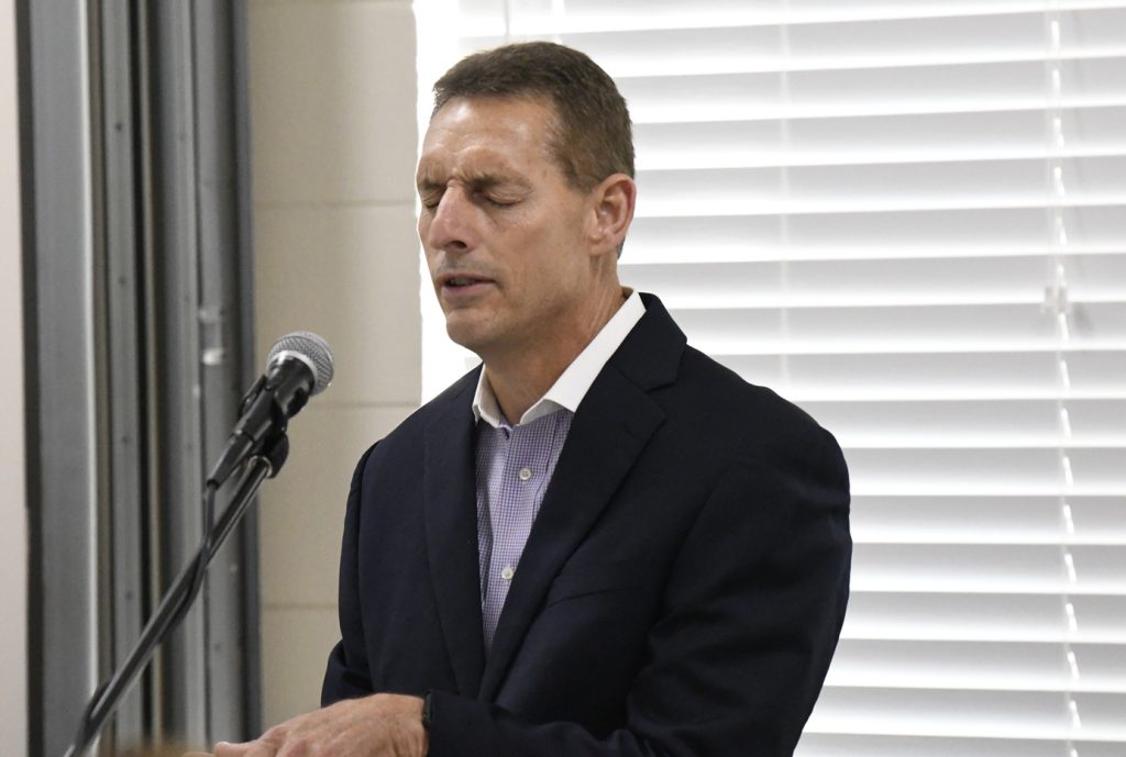 A man in a suit stands at a microphone with eyes closed, appearing to speak or sing, in front of white blinds.