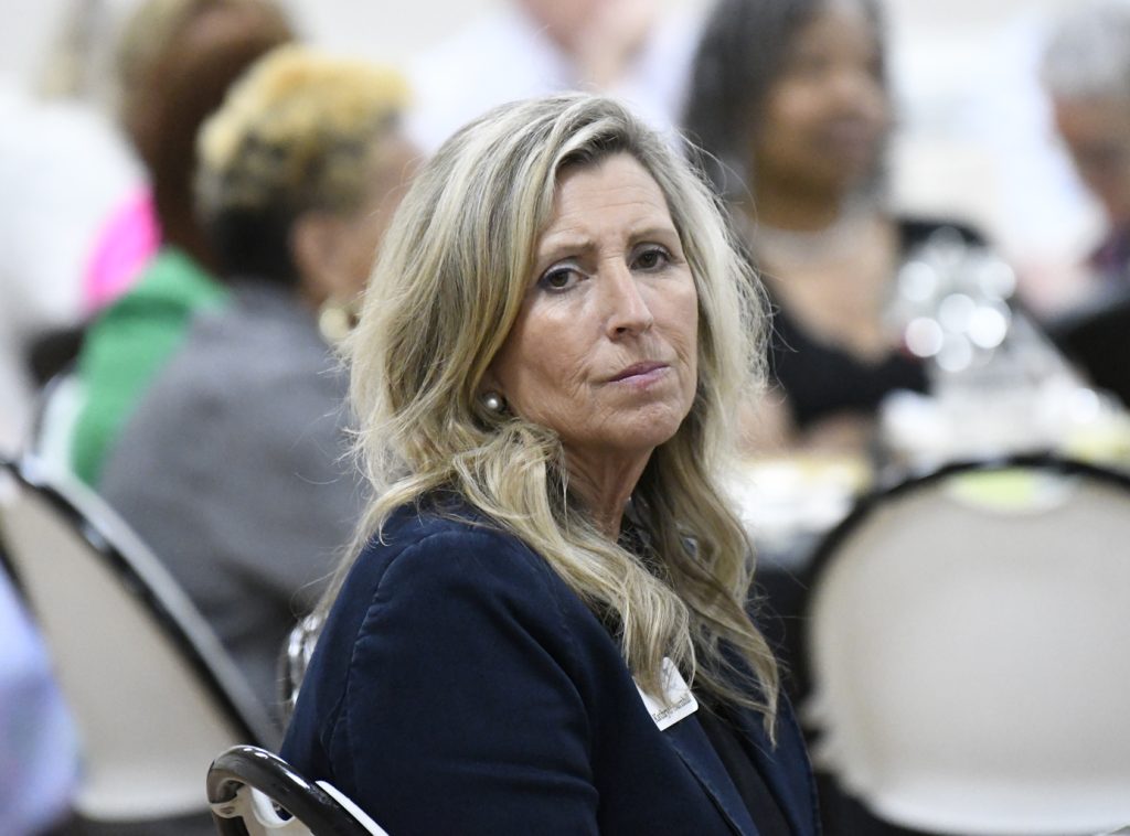 A woman with long blonde hair and a navy blazer sits at an event, looking at the camera with a serious expression. People are blurred in the background.
