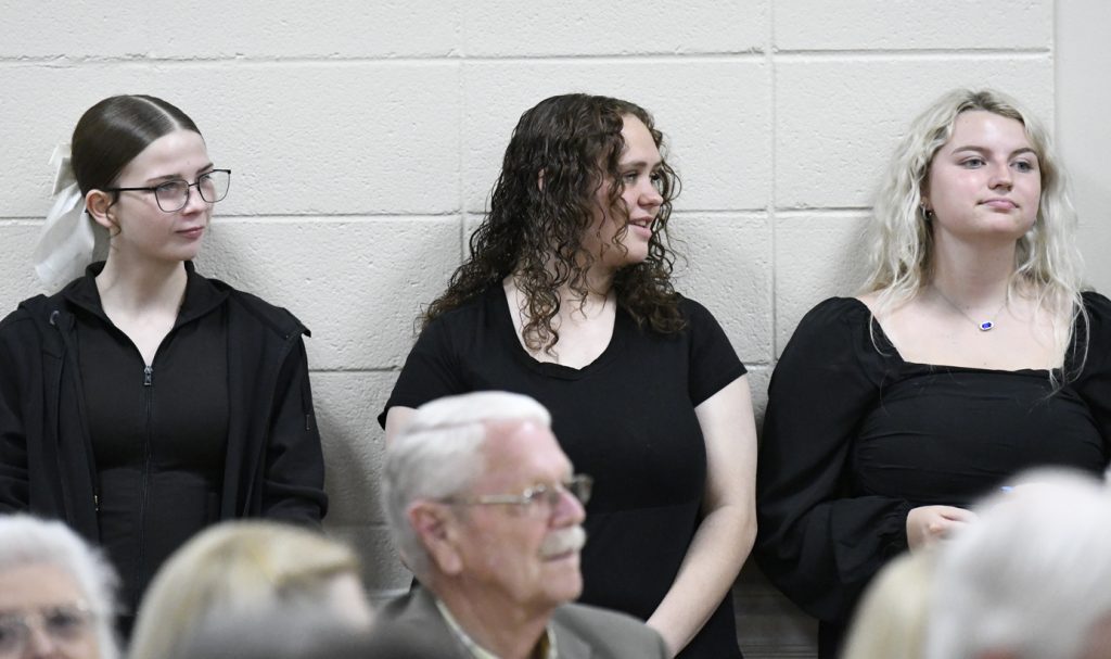 Three young women in black outfits stand against a white brick wall, observing something off-camera at a public indoor event with seated older adults in the foreground.