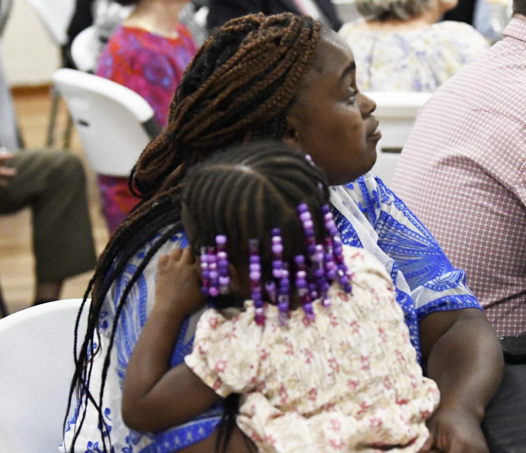 Woman in a blue patterned blouse holds a young girl with beaded braids, seated among others at an indoor event.