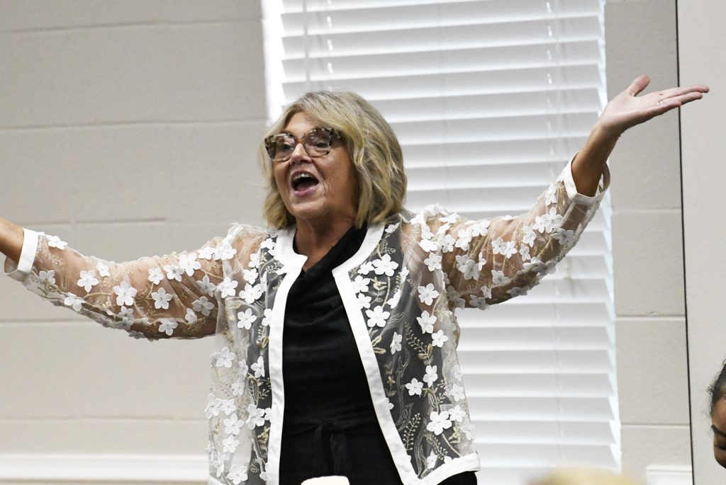 Woman with shoulder-length blonde hair, wearing glasses and a floral sheer jacket, stands indoors with arms raised, appearing to speak expressively.