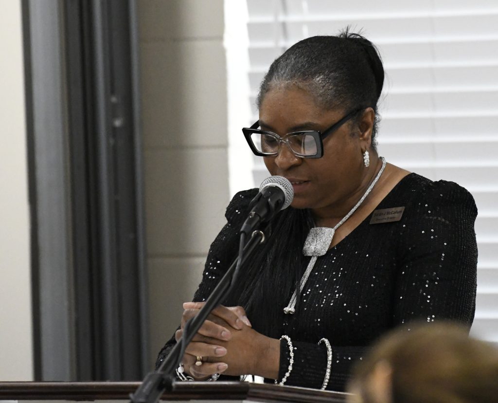 Woman in black sequin dress speaks into a microphone at a podium, wearing glasses and silver jewelry, standing in front of white blinds.
