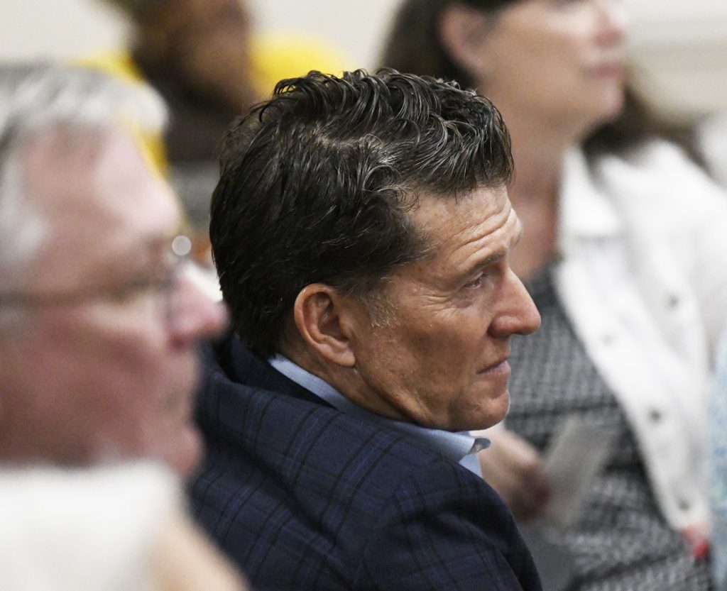 A man in a dark suit sits attentively among a blurred group of people at an indoor event or meeting.
