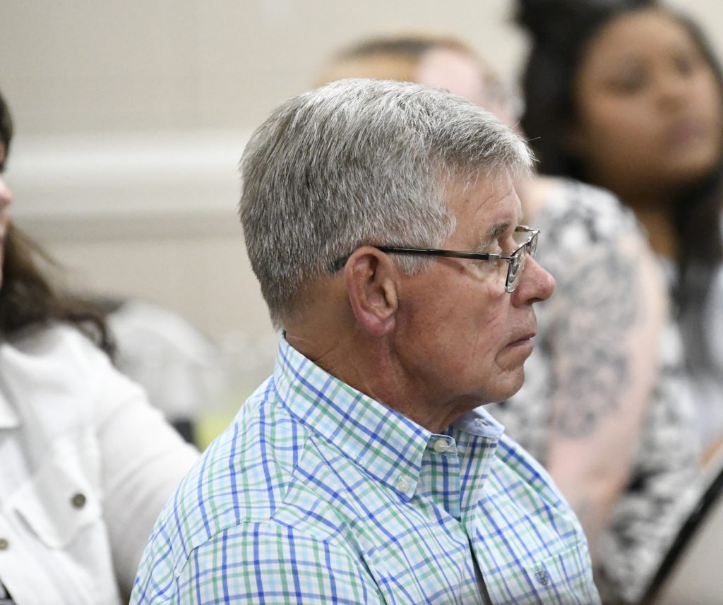 older man with gray hair and glasses wearing a plaid shirt sits and listens attentively in a group setting.