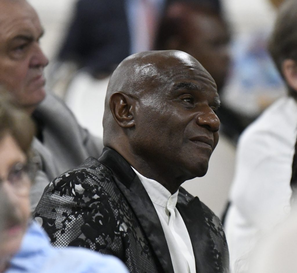 A man in a patterned suit jacket sits among a group of people, listening attentively during an indoor event or ceremony.