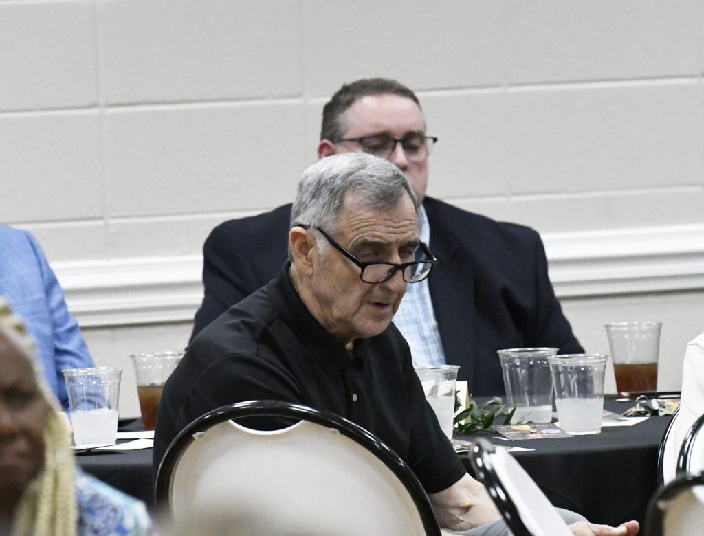 Two men sitting at a table during an indoor event, one wearing glasses and a black shirt, surrounded by empty cups and drinks.
