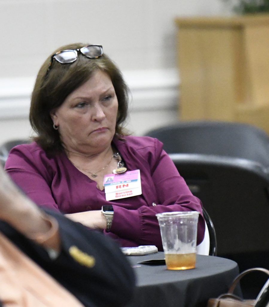 Woman with glasses on her head sits at a table looking serious, wearing a badge labeled "RN Nursing Director," with a half-full plastic cup in front of her.