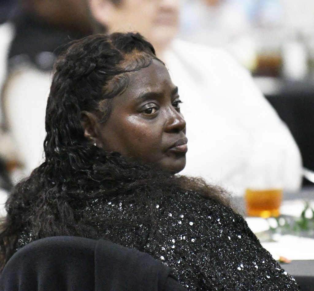 A woman with long curly hair and a sparkly black jacket sits at a table, looking to the side at a formal event.
