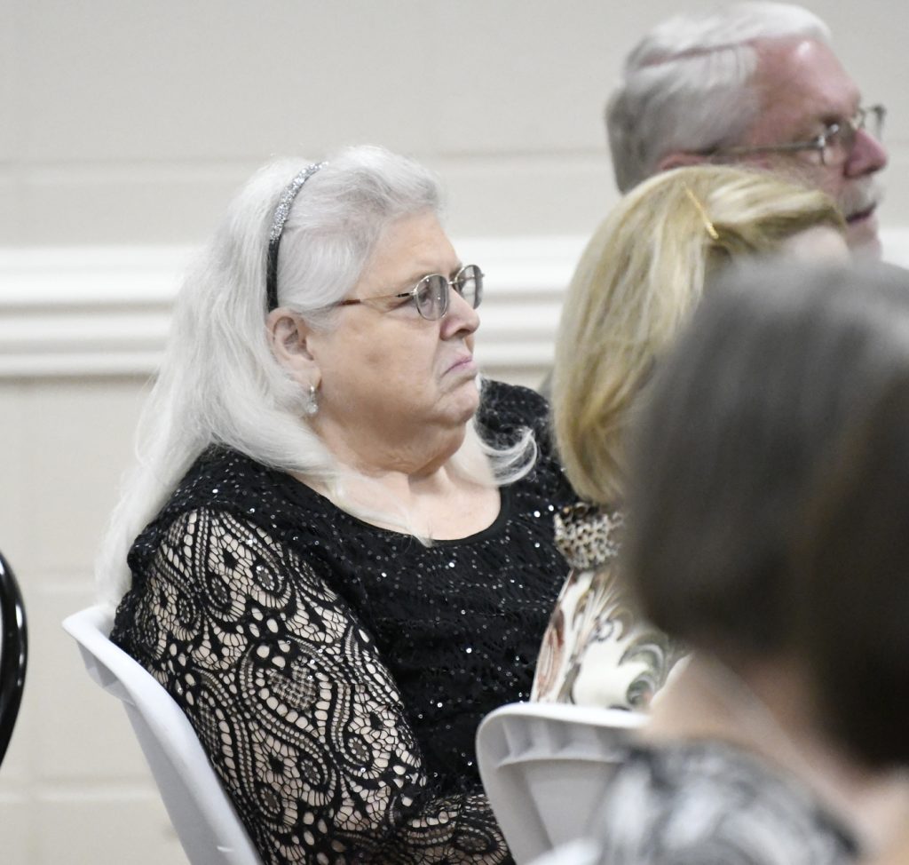Older woman with white hair and glasses sits in a crowd, wearing a black lace top and a headband, looking thoughtful or serious.