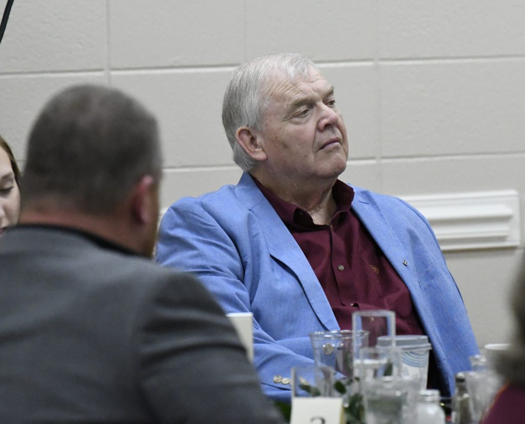 older man in a light blue blazer and burgundy shirt sits at a table, looking thoughtful during an indoor event.